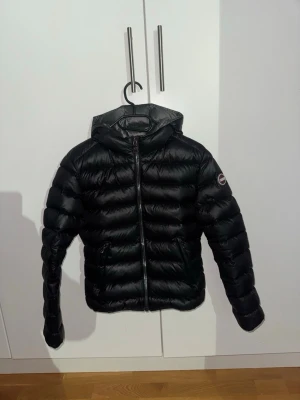 Svart Colmar pufferjacka med huva - Säljer en svart pufferjacka från Colmar med huva och dragkedja framtill. Jackan har en quiltad design och två fickor på sidorna. Perfekt för kalla dagar och riktigt snygg till streetwear-looken.