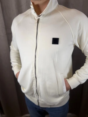 Vit ziptröja från Hugo Boss - Snygg vit ziptröja från Hugo Boss med hög krage och svarta detaljer. Tröjan har dragkedja hela vägen, två fickor och en svart logopatch på bröstet. Materialet är bomull och passformen är normal. Perfekt för en clean och stilren look.