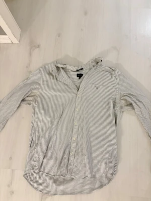 Randig skjorta från GANT XL - Snygg randig skjorta från GANT i storlek XL. Skjortan är vit med tunna blå ränder, har klassisk krage och knäppning framtill. Tillverkad i bomull!