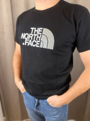 Svart t-shirt från The North Face - Svart t-shirt från The North Face med stor vit logga framtill. Klassisk rund hals och korta ärmar. Tillverkad i mjuk bomull som känns skön mot huden. Perfekt för dig som gillar streetwear och vill ha en clean look med tydlig branding.