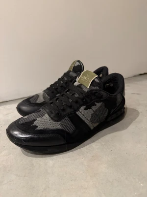 Valentino Rockrunner sneakers svart/grå - Säljer ett par Valentino Rockrunner sneakers i svart och grått med camouflage-mönster. Skorna har detaljer i mocka och skinn, samt nitar på hälen. Klassisk snörning och meshpaneler för extra cool look. Perfekta för dig som vill ha något unikt och stilrent. 41 men passar 42