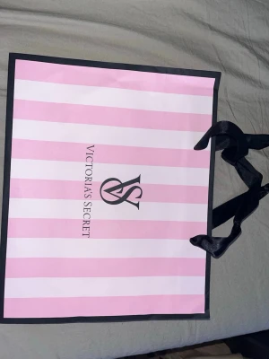 Randig shoppingväska Victoria's Secret - Säljer en ikonisk shoppingväska från Victoria's Secret med rosa och vita ränder och svarta detaljer. Väskan har svarta bandhandtag i satin och klassisk VS-logga på framsidan. Perfekt för dig som vill ha en snygg och praktisk påse till shopping eller förvaring.