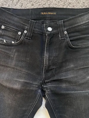 Svarta Nudie Jeans - Säljer ett par svarta jeans från Nudie Jeans med klassisk femficksdesign och snygga slitningar. Jeansen har smal passform, silverfärgade nitar och knapp, samt diskreta kontrastsömmar. Perfekta för dig som gillar en stilren och modern look. modell lean dean och längd 32