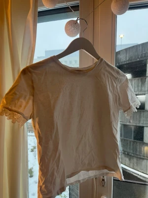 Vit topp med spetsdetalj från Zara - Säljer en vit topp från Zara i storlek S. T-shirten har korta ärmar med fina spetsdetaljer i ärmsluten och en klassisk rund halsringning. Materialet är mjukt och lätt, perfekt för en fräsch och enkel look.