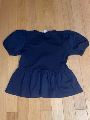 Mörkblå blus med puffärm och knyt - Supersöt mörkblå blus med puffiga korta ärmar och volang nedtill. Framtill har den tre dekorativa knytband som ger en extra detalj. Tillverkad i mjuk bomull, perfekt för dig som gillar en feminin och avslappnad stil.