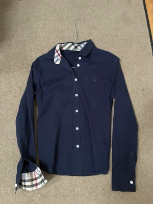 Mörkblå Burberry skjorta med rutig detalj - Snygg mörkblå skjorta från Burberry med klassiska vita knappar och ikoniskt rutigt mönster på insidan av kragen och manschetterna. Skjortan har lång ärm och en liten broderad logga på bröstet. Perfekt för dig som gillar stilrena och tidlösa plagg.