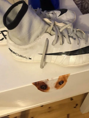 Nike MercurialX vita fotbollsskor - Säljer ett par vita Nike MercurialX fotbollsskor med högt skaft och svart Nike-logga. Skorna har snörning och är tillverkade i syntetmaterial med en tight passform. Perfekta för dig som vill ha stilrena och snabba skor på planen.