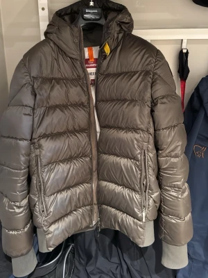 Grön Parajumpers jacka - Säljer en grön pufferjacka från Parajumpers med huva och dragkedja framtill. Jackan har quiltad design, ribbade muddar och en gul detalj vid halsen. Perfekt för kalla dagar och riktigt snygg till streetwear-stilen.