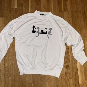 Vit sweatshirt med kattmotiv - Vit sweatshirt med rund hals och lång ärm. På bröstet finns ett broderat motiv av fyra katter i svart och grått. Tröjan har ribbade muddar vid ärmslut och nederkant, perfekt för dig som gillar gulliga detaljer.