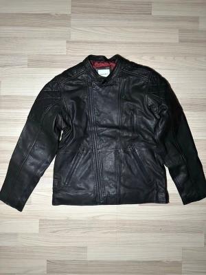 Svart skinnjacka från Vailent - Säljer en svart skinnjacka från Vailent med quiltade detaljer på axlar och nederkant. Jackan har flera dragkedjeförsedda fickor, långärmad design och ett vinrött, quiltat innerfoder. Snygg och tidlös modell som ger en cool look.