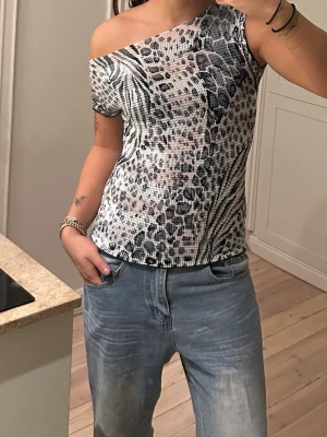 Leopardmönstrad topp från Miss Elegance - Säljer en snygg leopardmönstrad topp från Miss Elegance Essentials. Toppen är i svart, vit och grå ton, har en tight passform och är tillverkad i stretchigt syntetmaterial. Den har korta ärmar och kan bäras offshoulder för en trendig look.