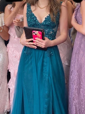 Blå balklänning med spets och paljetter - Säljer en blå balklänning med v-ringning där överdelen är dekorerad med blommig spets och glittriga paljetter, medan kjolen är i flera lager tyll och har en slits. Använd en gång, ordinarie pris 3400kr