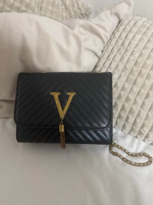 Svart quiltad axelväska med guldkedja - Säljer en svart quiltad axelväska från Valentino med guldfärgad kedja och stort V i metall framtill. Väskan har ett elegant mönster, dekorativ tofs och innerfack med dragkedja. Perfekt storlek för det viktigaste och riktigt snygg detalj till din outfit.