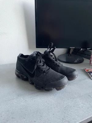 Nike Vapormax flyktig svarta sneakers strl 42 - Nike Vapormax sneakers i helsvart färg med stickad ovandel och transparent sula med luftkuddar. Skorna har snörning och en rund tå, samt diskret Nike-logga på sidan. Perfekt för dig som gillar sportig och modern stil.
