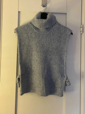 Ljusgrå stickad polotröja från Lindex - Supersnygg ljusgrå stickad polotröja från Lindex. Ärmlös modell med bred polokrage och ribbad struktur. Sidorna har fina knytband som ger en extra detalj. Perfekt att slänga över en skjorta eller långärmad topp.