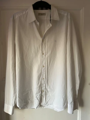 Vit skjorta från Burberry London - Vit skjorta från Burberry London i klassisk design med broderad logga på bröstet. Skjortan har lång ärm, knappar framtill och är tillverkad i mjuk bomull. Perfekt för dig som gillar stilrena och tidlösa plagg med exklusiv känsla.