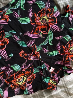 Blommig skjorta från Levi's i svart - Säljer en svart skjorta från Levi's i storlek L med stora, färgglada blommor i rött, gult, rosa och grönt. Skjortan har klassisk krage, knappar framtill och en bröstficka. Materialet är mjuk bomull som känns skönt mot huden. Perfekt för dig som gillar att sticka ut med mönster!