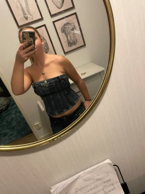 Mörkblå denim tubtopp - Säljer en snygg mörkblå tubtopp i denim med markerade sömmar och knappdetalj framtill. Toppen är axelbandslös och har en tight passform som framhäver midjan. Perfekt att styla med jeans eller kjol för en cool look.