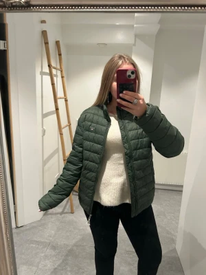 Grön jacka  från GANT - Snygg mörkgrön pufferjacka från Tommy Hilfiger med quiltad design och dragkedja framtill. Jackan har en klassisk passform och är perfekt för kyligare dagar. Liten logga på bröstet och diskret, stilren look som funkar till det mesta.