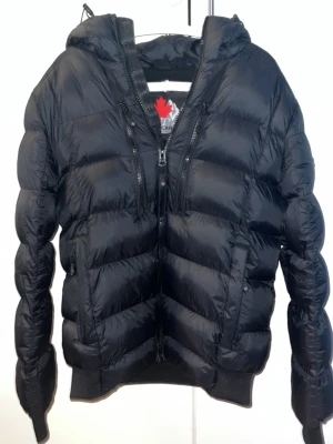 Svart pufferjacka från Zavetti Canada - Säljer en svart pufferjacka från Zavetti Canada med huva och feta detaljer som broderad lönnlövspatch på ärmen och stor logga på ryggen. Jackan har flera dragkedjor framtill och är riktigt varm, perfekt för kalla dagar. Materialet är syntet och jackan har en schysst, normal passform.