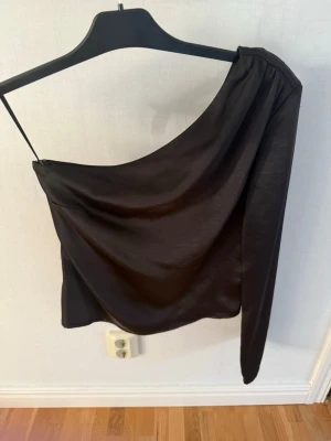 Svart oneshoulder topp Gina Tricot - Säljer en svart oneshoulder topp från Gina Tricot i storlek 34. Materialet är glansigt och känns mjukt, vilket ger en snygg och modern look. Fint skick 
