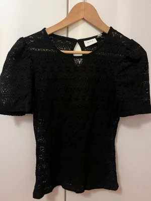 Svart spetsblus från Vila - Supersnygg svart blus från Vila i spets med korta puffärmar och rund halsringning. Blusen har ett fint hålmönster och knapp i nacken och lite genomskinlig. Aldrig använd!!❤️