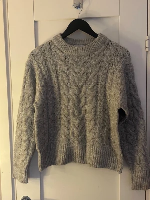 Grå stickad tröja från Gina Tricot - Mysig grå stickad tröja från Gina Tricot med grovt flätmönster och ribbade muddar. Perfekt för kyliga dagar med sin höga hals och långa ärmar. Passar dig som gillar en klassisk och enkel look. Storlek 146/152 men passar en XS/S