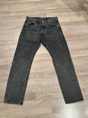 Levi's 501 svarta jeans W29 L30 - Svarta Levi's 501 jeans med klassisk rak passform och fem fickor. Jeansen har en snygg tvättad look och är tillverkade i slitstarkt bomullsmaterial. Ikonisk läderpatch bak i midjan och röd Levi's-tag på bakfickan. Perfekta för dig som gillar tidlös stil.