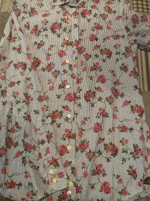 Randig blommig skjorta från Morris - Säljer en skjorta från Morris i storlek S med blåvita ränder och blommigt mönster i rött och grönt. Skjortan har långa ärmar, klassisk krage och knäppning framtill. Perfekt för dig som vill sticka ut med en färgglad och unik look.