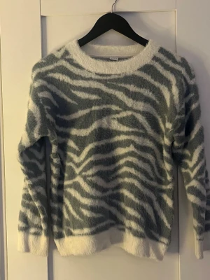 Zebramönstrad fluffig tröja - Supermjuk och fluffig långärmad tröja med zebramönster i grått och vitt. Rund hals och breda muddar vid ärmslut och nederkant. Perfekt för dig som gillar djurmönster och vill ha något riktigt cozy till hösten och vintern.