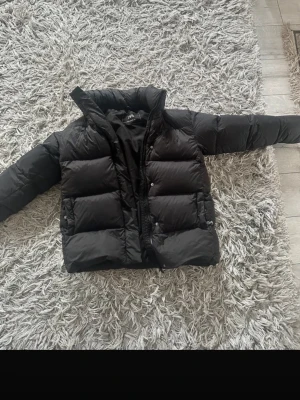 Svart pufferjacka från ZARA XS - Säljer en svart pufferjacka från ZARA i storlek XS. Jackan har hög krage, tryckknappar och dragkedja framtill. Den är vadderad och har en klassisk, boxig passform som är perfekt för kalla dagar. Vinter jacka