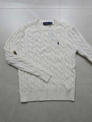 Vit stickad tröja från Polo Ralph Lauren - Säljer en vit stickad tröja från Polo Ralph Lauren med klassiskt kabelstickat mönster och rund hals. Tröjan har långärmade ärmar och en liten broderad logga i marinblått på bröstet. Perfekt för dig som gillar stilrena och tidlösa plagg.