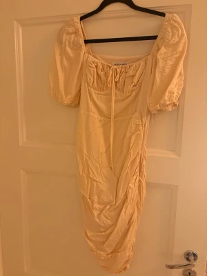 Ljus beige långklänning från Adoore, Positano  - Supersnygg ljus beige långklänning från Adoore med puffärmar, snörning vid bysten och draperad, rynkad nederdel. Klänningen har en romantisk vibe och är tillverkad i ett mjukt, glansigt material som känns lyxigt.