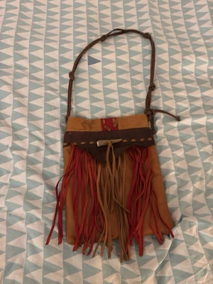 Brun axelväska med fransar - Säljer en unik brun axelväska med långa fransar i rött och beige. Väskan har justerbar axelrem med knutar och dekorativt band upptill. Perfekt för dig som gillar boho-stil och vill sticka ut med coola detaljer.
