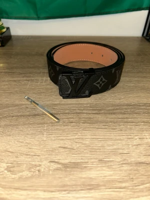 L&V bälte - Snyggt bälte med klassiskt rutigt mönster i svart och grått. Bältet har en stor logga i svart metall som spänne och insidan är i ljusbrunt skinn. Perfekt för att lyfta din outfit med en lyxig touch.