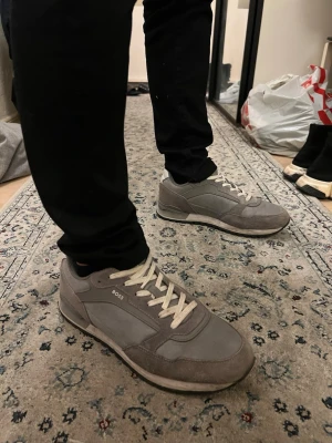 Grå sneakers från BOSS med snörning - Säljer ett par stilrena grå sneakers från BOSS med vit snörning och platt sula. Skorna är tillverkade i en kombination av mocka och syntetmaterial. Perfekta för dig som gillar en clean och modern look.