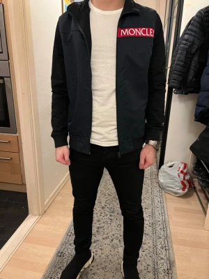 Svart Moncler bomberjacka med röd logga - Säljer en svart bomberjacka från Moncler med stor röd logga på bröstet och emblem på ärmen. Jackan har dragkedja framtill, ribbade muddar och en clean, sportig look. Materialet är polyester och bomull, perfekt för dig som gillar stilrena och exklusiva plagg.