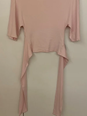 Ljusrosa topp med knyt - Säljer en ljusrosa omlottopp med knyt framtill. Toppen har korta ärmar och är gjord i mjukt bomullsmaterial. Den har en croppad passform och långa band som kan knytas i midjan för en snygg detalj. Perfekt för att skapa en chill och trendig look.