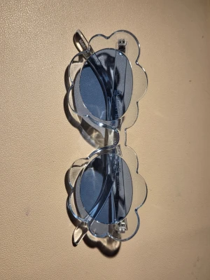  solglasögon - Säljer ett par unika solglasögon med transparent, klar plastbåge och runda glas i grå ton. Glasögonen har en cool blomformad design runt glasen som verkligen sticker ut. Perfekta för dig som vill ha något lekfullt och trendigt till sommaren!