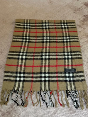 Rutig ullväst från Burberrys - Säljer en klassisk rutig halsduk från Burberrys i mjuk ull. Halsduken har beige bas med svarta, vita och röda rutor, samt fransar i ändarna. Perfekt för dig som gillar ikoniska mönster och vill ha något som sticker ut.