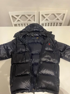 Mörkblå pufferjacka från Polo Ralph Lauren - Snygg mörkblå pufferjacka från Polo Ralph Lauren med huva och två sidofickor med dragkedja. Jackan har RL-detaljer på dragkedjorna och ett rött Polo-logo på bröstet. Materialet är glansigt och känns riktigt mjukt, perfekt för kalla dagar. Pris kan diskuteras!