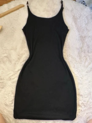 Svart tight kortklänning - Säljer en svart, figurnära kortklänning med smala axelband. Klänningen har en enkel och stilren design som framhäver siluetten. Perfekt för dig som gillar minimalistisk stil och vill ha en klassisk svart klänning i garderoben.