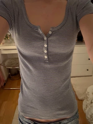 Randig blåvit topp med knappar från brandy melville  - Säljer en slim, kortärmad topp med blåvita ränder och rundad halsringning. Toppen har fem vita knappar framtill och är gjord i mjuk bomull. Perfekt till jeans och enkel att styla för olika tillfällen. Storleken är XS