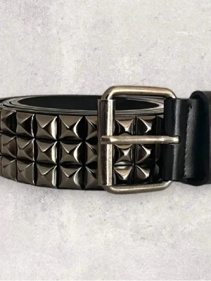 Svart Emo välte med nitar  - Säljer ett svart bälte i skinn med silvriga fyrkantiga nitar längs hela bandet. Bältet har ett metallspänne och är riktigt chunky, perfekt för att ge din outfit en edgy vibe. Passar till jeans eller över en kavaj för att bryta av stilen.