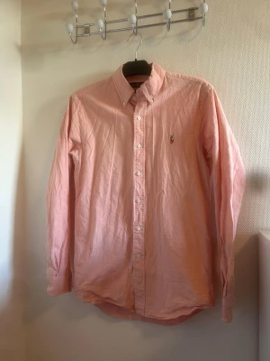 Rosa skjorta från Ralph Lauren  - Säljer en snygg rosa skjorta från Ralph Lauren i slim fit. Skjortan har klassisk button-down krage, vita knappar och det ikoniska broderade logot på bröstet. Perfekt för dig som gillar stilrena och fräscha plagg. Hör av dig vid minsta lilla fråga!