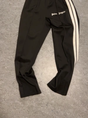 Palm Angels svarta trackpants med vita ränder - Svarta trackpants från Palm Angels med två vita ränder längs sidorna och logotypen tryckt på höften. Byxorna har elastisk midja och är gjorda i ett glansigt syntetmaterial som ger en sportig vibe. Perfekt för dig som gillar streetwear och vill sticka ut.