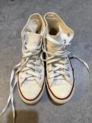 Converse All Star vita high tops 37.5 - Säljer ett par klassiska Converse All Star high tops i vit textil med röd kant och rund tå. Skorna har snörning och platt sula, samt den ikoniska loggan på sidan. Perfekt för dig som gillar retrostil och vill ha ett par tidlösa sneakers.