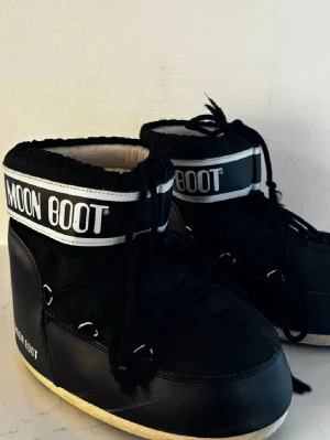 Svarta Moon Boots med snörning - Säljer ett par svarta Moon Boots med vit logga och snörning framtill. Skorna har en rund tå och platt sula, perfekt för kalla vinterdagar. Materialet är syntet och de har en klassisk moon boot-design med högt skaft.