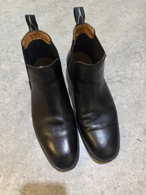 Svarta chelsea boots från Pace, strl 42 - Säljer ett par stilrena svarta chelsea boots från Pace i storlek 42. Skorna är gjorda i skinn med elastiska paneler på sidorna och har en rund tå. Insidan är fodrad i brunt tyg och de har en klassisk platt sula. Perfekta för dig som gillar enkel och tidlös design.