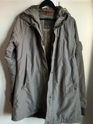 Grå parkajacka från Didriksons, strl 38 - Säljer en grå parkajacka från Didriksons med quiltat foder och huva. Jackan har gul dragkedja, praktiska fickor och snygga detaljer. Perfekt för kyliga dagar och har en längre passform som ger extra skydd mot väder och vind.